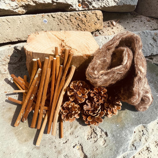 Natural bug hotel habitat starter kit components including dried pine needles, pine cones, and a rolled piece of brown felt wool, arranged on a stone surface next to a cut wood log.